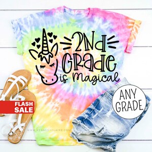 Op de afbeelding: Een tie-dye T-shirt met een eenhoorn en de tekst "2nd Grade is magical". Het shirt is roze, geel, blauw en groen.