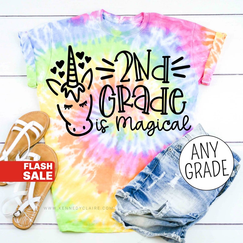 Op de afbeelding: Een tie-dye T-shirt met een eenhoorn en de tekst "2nd Grade is magical". Het shirt is roze, geel, blauw en groen.