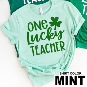 May include: Mint green t-shirt with the text "ONE LUCKY TEACHER" in green, along with a green shamrock. The shirt is a light, pastel color, perfect for St. Patrick's Day or any day to celebrate teachers.