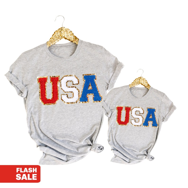 May include: Two gray t-shirts, one adult and one child size, each displaying the word "USA" in red, white, and blue sequined letters. The shirts are hanging on gold glitter hangers against a white background. A red "FLASH SALE" banner is in the lower left corner.