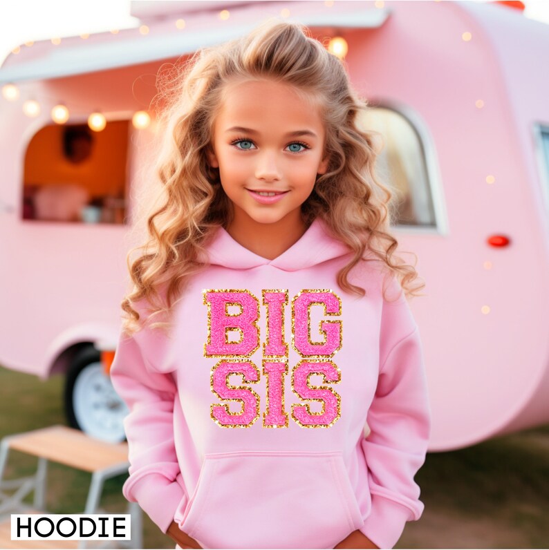 May include: A pink hoodie with a gold glitter design that says "BIG SIS" on the front. The hoodie is being worn by a young girl with long blonde hair.