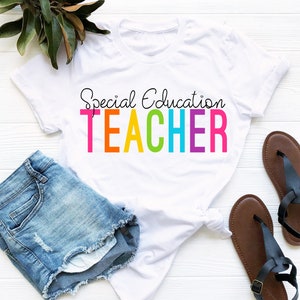May include: White t-shirt with the text "Special Education TEACHER" in rainbow colors. The shirt is laying on a white surface with a pair of blue denim shorts and brown sandals.