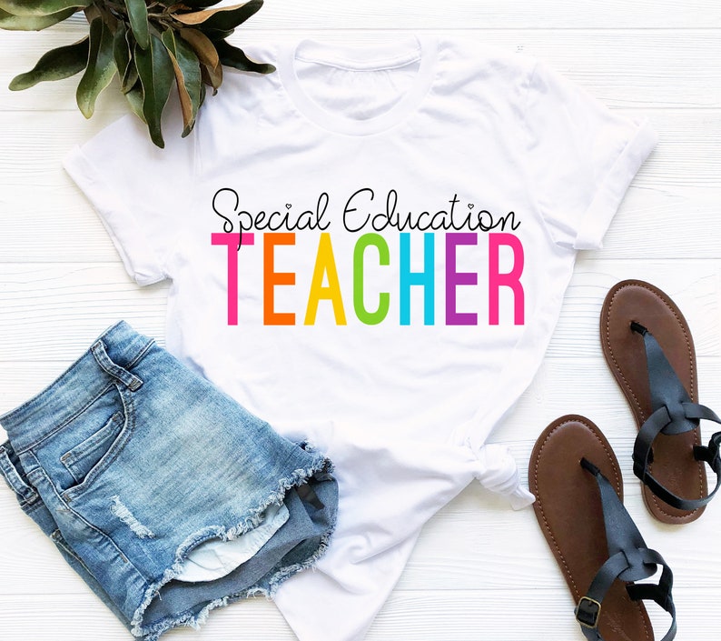 May include: White t-shirt with the text "Special Education TEACHER" in rainbow colors. The shirt is laying on a white surface with a pair of blue denim shorts and brown sandals.