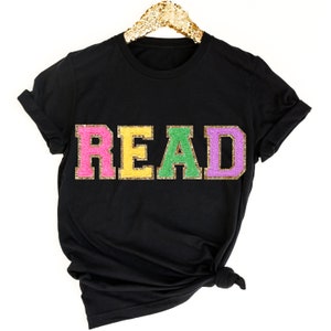 May include: Black t-shirt with the word "READ" in colorful glitter letters. The letters are pink, yellow, green, and purple.