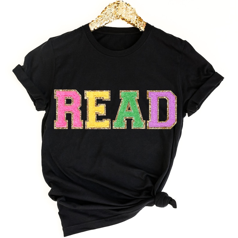 May include: Black t-shirt with the word "READ" in colorful glitter letters. The letters are pink, yellow, green, and purple.