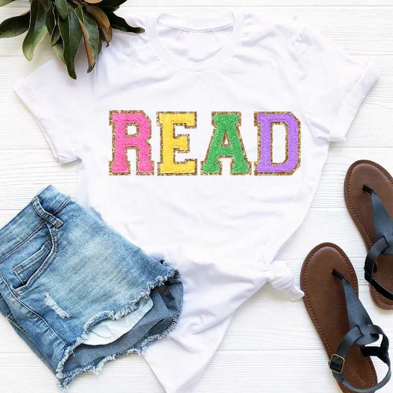 May include: White t-shirt with the word "READ" in colorful glitter letters. The letters are pink, yellow, green, and purple. The shirt is paired with denim shorts and brown sandals.