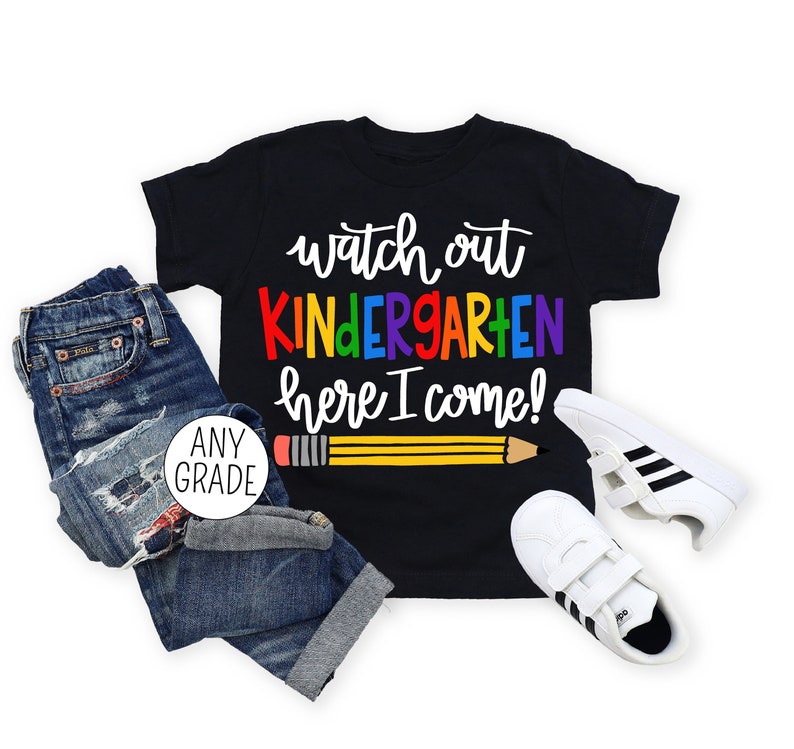 May include: Black t-shirt with the text "Watch out Kindergarten here I come!" in rainbow colours with a yellow pencil underneath. The shirt is on a white background with a pair of white trainers with black stripes and a pair of blue jeans with a white circle that says "Any Grade".