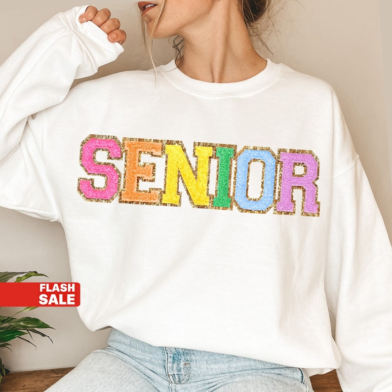 May include: White sweatshirt with the word "SENIOR" in rainbow letters with gold glitter accents.