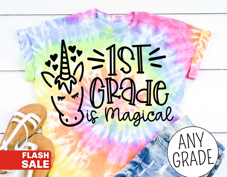Op de afbeelding: Een tie-dye T-shirt met een eenhoorn en de tekst "1st Grade is magical".