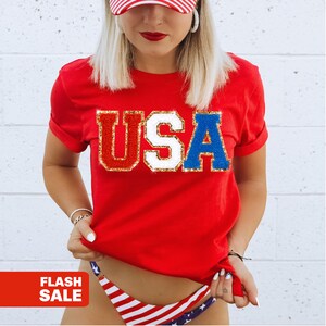 May include: A red t-shirt with the word "USA" in a sequined design. The letters are in red, white, and blue with gold trim. The image also includes a red and white striped baseball cap and a bikini bottom with a USA flag design.