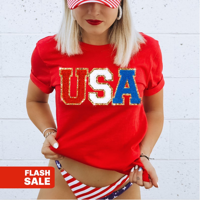 May include: A red t-shirt with the word "USA" in a sequined design. The letters are in red, white, and blue with gold trim. The image also includes a red and white striped baseball cap and a bikini bottom with a USA flag design.