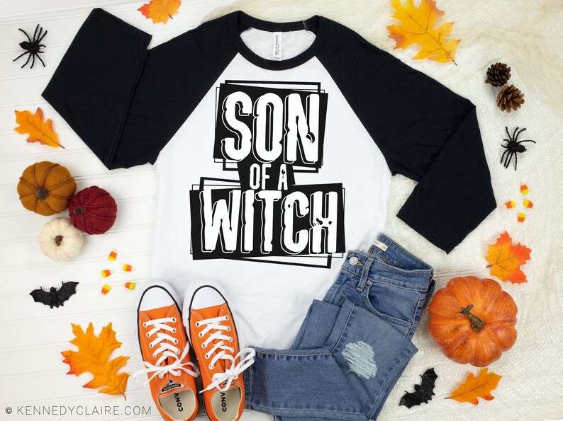 以下が含まれることがあります： 大胆なブロック体のフォントで「SON OF A WITCH」の文字が入った、白と黒のラグランスタイルのシャツ。オレンジ色のスニーカー、ブルージーンズ、カボチャ、葉、クモなどのハロウィーンの装飾が施された画像です。