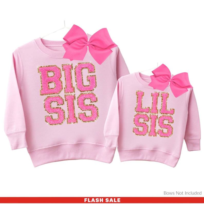 May include: Two pink long-sleeved shirts with gold glitter lettering. The larger shirt says "BIG SIS" and the smaller shirt says "LIL SIS".