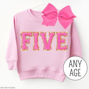 May include: A light pink sweatshirt with the word "FIVE" in pink and gold sequined letters. A large pink bow is attached to the top. The text "ANY AGE" is in a white circle.