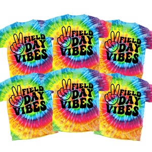 Puede incluir: Seis camisetas te&ntilde;idas con la inscripci&oacute;n "Field Day Vibes" en escritura negra y un gr&aacute;fico de signo de la paz. Las camisetas tienen un patr&oacute;n de remolino arco&iacute;ris de azul, amarillo, rosa y naranja.