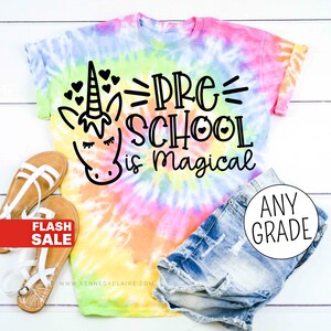 Op de afbeelding: Een tie-dye T-shirt met een eenhoorn en de tekst "Pre-School is magical". Het shirt ligt op een wit houten oppervlak met een paar sandalen en een spijkerbroek.