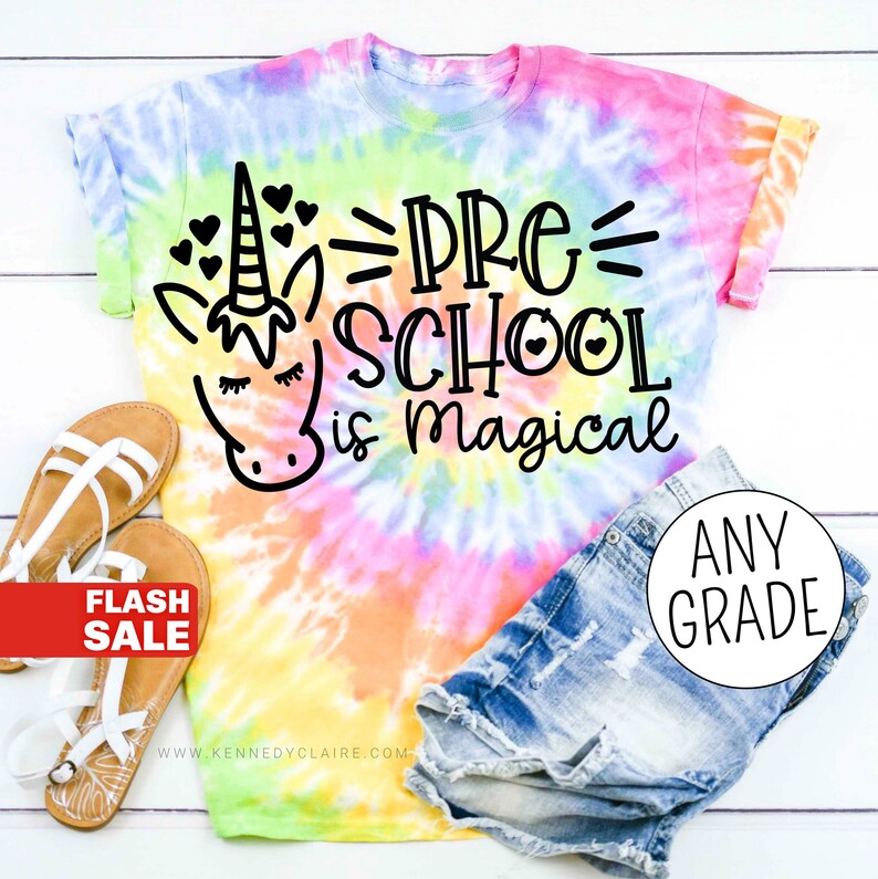 Op de afbeelding: Een tie-dye T-shirt met een eenhoorn en de tekst "Pre-School is magical". Het shirt ligt op een wit houten oppervlak met een paar sandalen en een spijkerbroek.