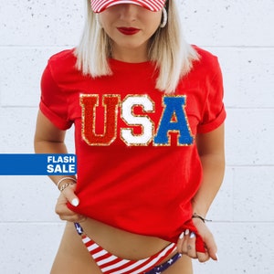 May include: Red, white, and blue t-shirt with the word "USA" in glitter letters. The shirt is being worn by a person with a red, white, and blue hat.
