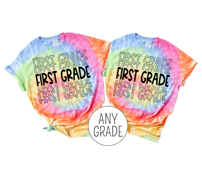 Pu&ograve; includere: Due magliette tie-dye con il testo "FIRST GRADE" ripetuto in un motivo arcobaleno. Le magliette sono di colore blu chiaro e giallo con un motivo tie-dye arcobaleno. Il testo &egrave; in nero.