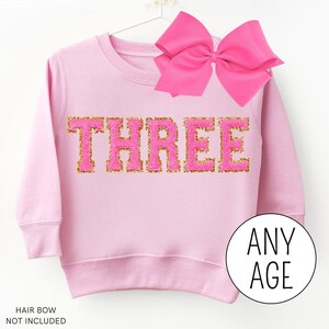 May include: Pink sweatshirt with gold glitter lettering that says "THREE". The sweatshirt is for a child and has a pink bow on the top.