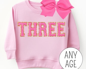 May include: Pink sweatshirt with gold glitter lettering that says "THREE". The sweatshirt is for a child and has a pink bow on the top.
