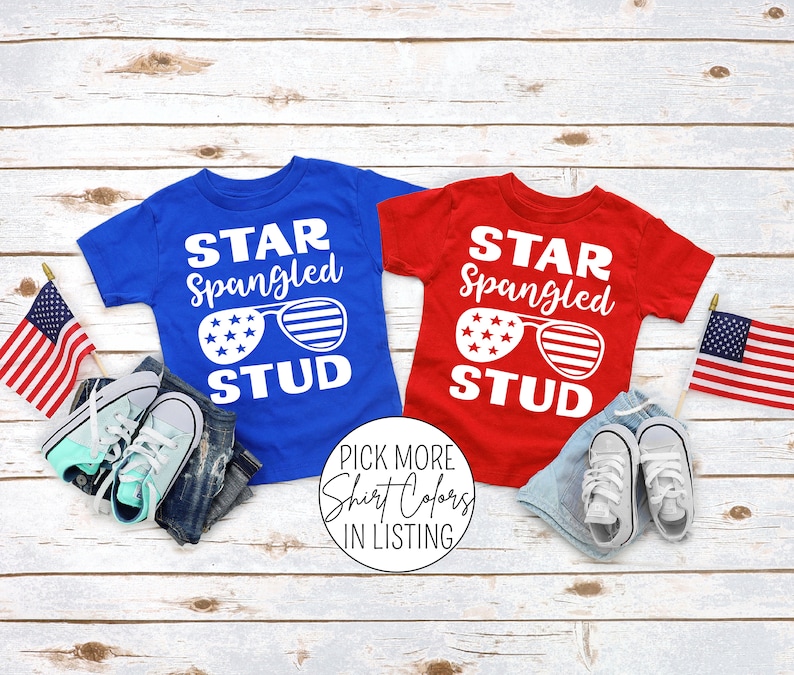 May include: Two red and blue t-shirts with the text "Star Spangled Stud" and a graphic of sunglasses with stars and stripes. The shirts are laid out on a white wood background with American flags and sneakers.