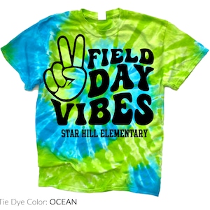 Puede incluir: Una camiseta te&ntilde;ida con tinte anudado en tonos azules y verdes. La camiseta presenta las palabras "FIELD DAY VIBES" en negro, un signo de la paz y el texto "STAR HILL ELEMENTARY". El dise&ntilde;o de la camiseta es brillante y colorido.