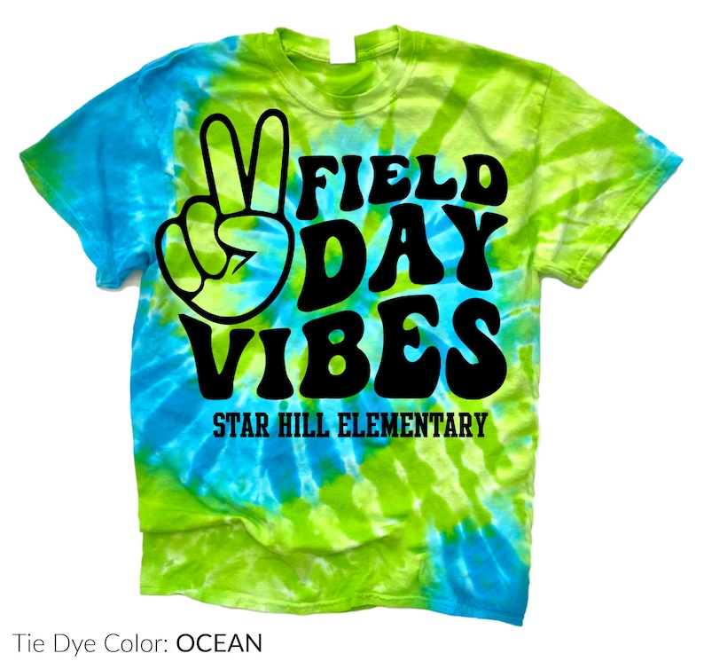 Puede incluir: Una camiseta te&ntilde;ida con tinte anudado en tonos azules y verdes. La camiseta presenta las palabras "FIELD DAY VIBES" en negro, un signo de la paz y el texto "STAR HILL ELEMENTARY". El dise&ntilde;o de la camiseta es brillante y colorido.