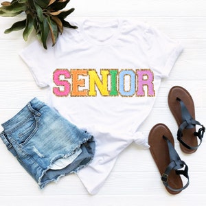 May include: White t-shirt with the word "SENIOR" in rainbow glitter letters. The shirt is paired with denim shorts and brown sandals.