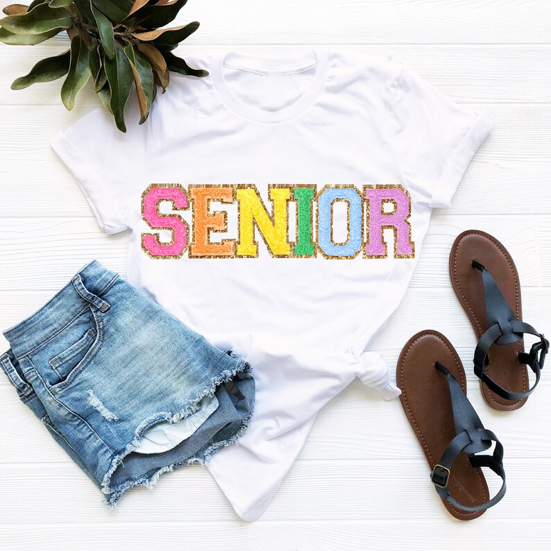 May include: White t-shirt with the word "SENIOR" in rainbow glitter letters. The shirt is paired with denim shorts and brown sandals.