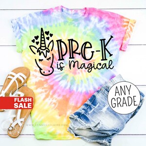 Op de afbeelding: Een tie-dye T-shirt met een eenhoorn en de tekst "Pre-K is magical". Het shirt ligt op een wit houten oppervlak met een paar sandalen en een spijkerbroek.