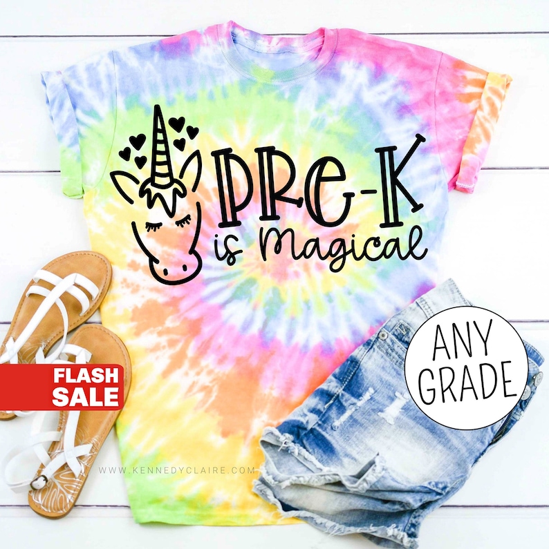 Op de afbeelding: Een tie-dye T-shirt met een eenhoorn en de tekst "Pre-K is magical". Het shirt ligt op een wit houten oppervlak met een paar sandalen en een spijkerbroek.