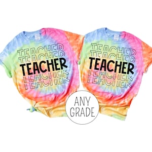 Pu&ograve; includere: Due magliette tie-dye con il testo "Teacher" ripetuto in uno schema impilato. Le magliette sono in tonalit&agrave; di rosa, arancio, giallo e blu. Il testo &egrave; nero. Le magliette sono etichettate "Any Grade".