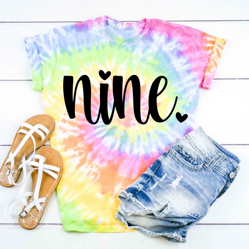 May include: A tie-dye t-shirt with the word "nine" in black script with a small heart above and below the word. The shirt is paired with denim shorts and white sandals.