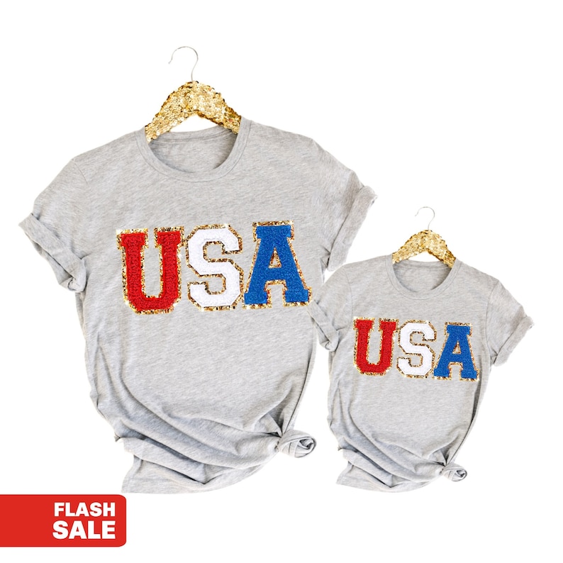May include: Two grey t-shirts with the word "USA" in red, white, and blue sequins on a white background.