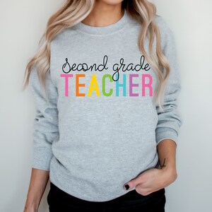 Puede incluir: Una sudadera gris con el texto "Second grade TEACHER" en colores arco&iacute;ris.