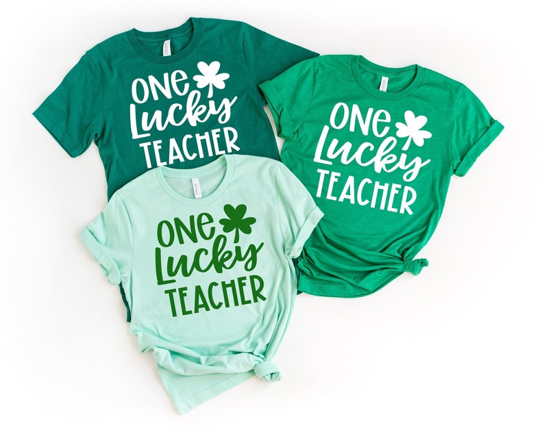 May include: Three green t-shirts with the text "ONE Lucky TEACHER" and a shamrock graphic. The shirts are in shades of emerald, mint, and kelly green. The text is in a white, bold, sans-serif font.