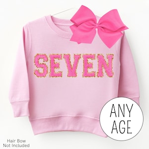 May include: Pink sweatshirt with the word "SEVEN" in pink and gold glitter letters. The sweatshirt is on a hanger and has a pink bow on top.