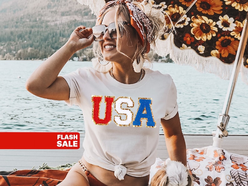 May include: A cream-colored t-shirt with the word "USA" in red, white, and blue sequined letters. The image shows a person wearing sunglasses and a patterned headscarf, with a red banner that says "FLASH SALE".