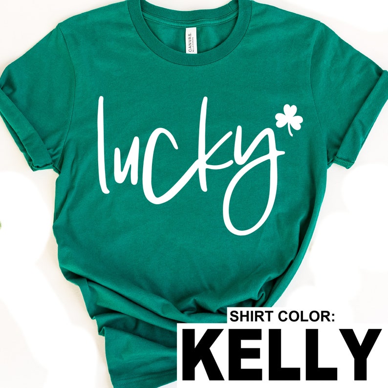 May include: Kelly green t-shirt with the word "lucky" in white script and a white shamrock. The shirt has rolled-up sleeves and a crew neck. The text "SHIRT COLOR: KELLY" is at the bottom of the image.