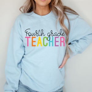 Puede incluir: Sudadera azul claro con el texto "fourth grade TEACHER" en colores arco&iacute;ris.