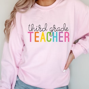 Puede incluir: Sudadera rosa con el texto "third grade TEACHER" en colores arco&iacute;ris.