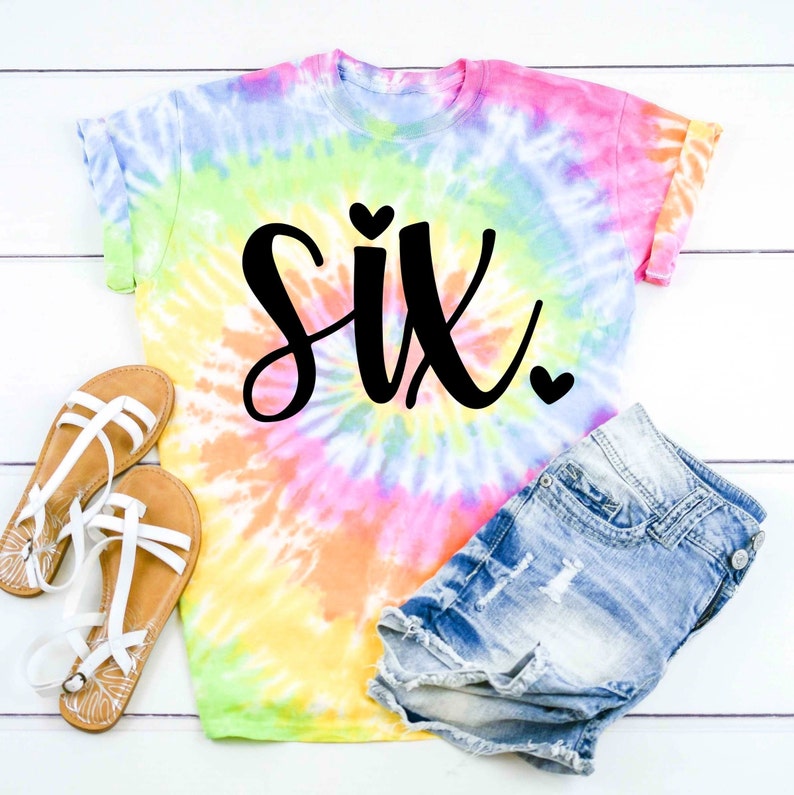May include: A tie-dye t-shirt with the word "six" in black script with two small hearts. The shirt is on a white wooden surface with a pair of white sandals and denim shorts.