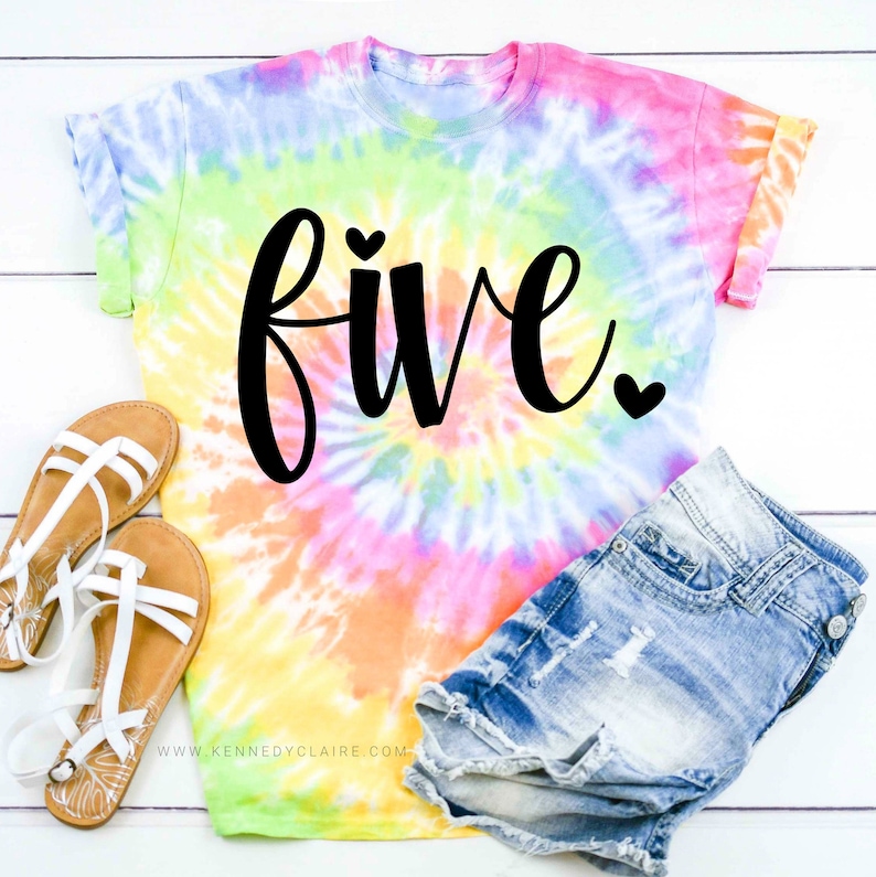 May include: A tie-dye t-shirt with the word "five" in black script and a small heart on each side of the word. The shirt is on a white wooden surface with a pair of sandals and denim shorts.