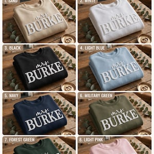 May include: A collection of eight crewneck sweatshirts in sand, white, black, light blue, navy, military green, forest green, and light pink. Each sweatshirt features the text "mrs. BURKE" in raised, white, dimensional lettering across the chest.