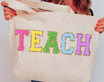 Puede incluir: Una bolsa de lona blanca con la palabra "TEACH" escrita en letras coloridas y brillantes. Las letras son rosa, amarillo, verde, azul claro y morado.