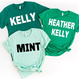 May include: Three short-sleeved t-shirts in shades of green and mint. The shirts have white text that reads "KELLY", "HEATHER KELLY", and "MINT". The shirts are laid flat on a white surface.