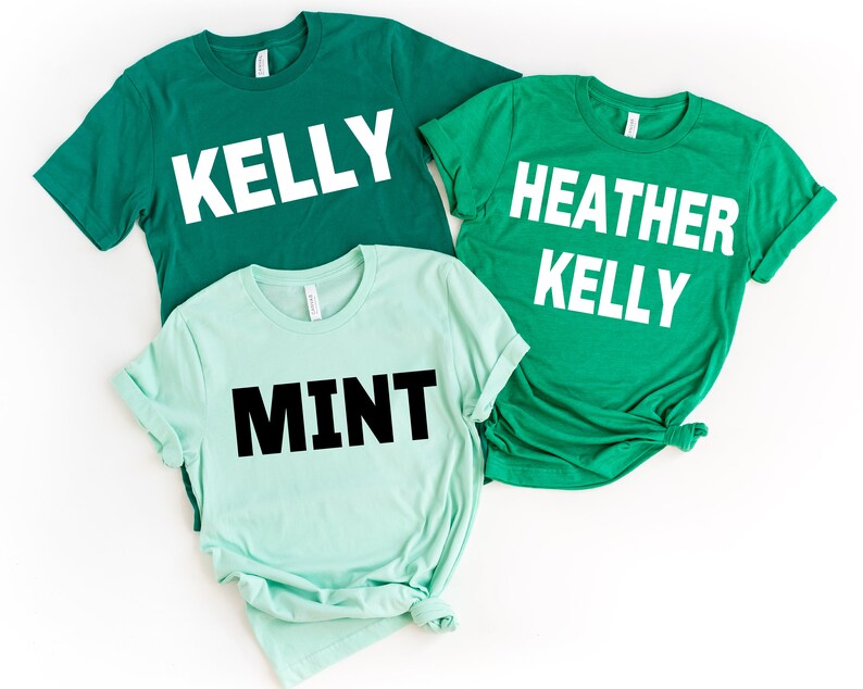 May include: Three short-sleeved t-shirts in shades of green and mint. The shirts have white text that reads "KELLY", "HEATHER KELLY", and "MINT". The shirts are laid flat on a white surface.
