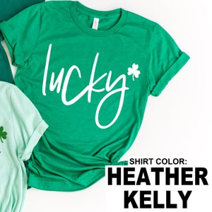 May include: A green t-shirt with the word "lucky" in white script and a white shamrock. The shirt is a heather kelly color. Another green shirt is partially visible in the background.