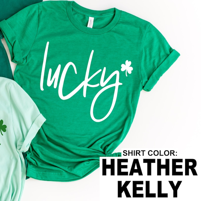 May include: A green t-shirt with the word "lucky" in white script and a white shamrock. The shirt is a heather kelly color. Another green shirt is partially visible in the background.